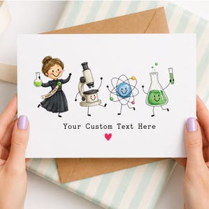Può includere: Un biglietto d'auguri bianco con un'illustrazione a cartoni animati di Marie Curie, un microscopio, un atomo e un becher con liquido verde, tutti con facce sorridenti. Sotto, il testo Your Custom Text Here con un piccolo cuore rosso.