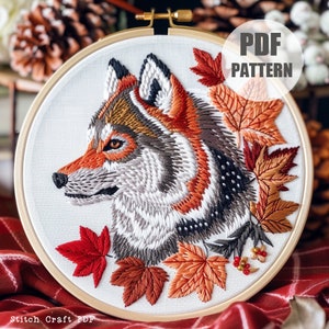 Może przedstawiać: Haftowany obraz w obręczy przedstawiający szczegółowy portret głowy wilka z wzorem futra w kolorze szarym, białym i pomarańczowym. Wilk jest otoczony jesiennymi liśćmi w odcieniach pomarańczowego, czerwonego i brązowego.