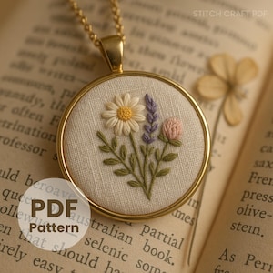 Może przedstawiać: Złoty naszyjnik z okrągłym, haftowanym wzorem kwiatowym. Haft przedstawia stokrotkę, lawendę i różowy kwiat z zielonymi liśćmi. Wisiorek jest umieszczony na kremowej tkaninie i oznaczony napisem "STITCH CRAFT PDF".