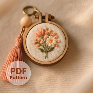 Może przedstawiać: Drewniany brelok do haftu z kwiatowym wzorem. Haft przedstawia brzoskwiniowe i białe kwiaty z zielonymi liśćmi. Brzoskwiniowy frędzel i drewniana kulka wiszą u góry. Widoczny jest tekst "PDF Pattern".