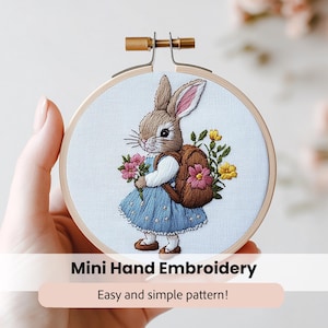 May include: A hand embroidery hoop with a cute bunny wearing a blue dress and a brown backpack. The bunny is holding a bouquet of pink and yellow flowers. The hoop is made of wood and has a white fabric background. The text "Mini Hand Embroidery" and "Easy and simple pattern!" is below the hoop.