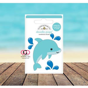 May include: A cardstock sticker featuring a light blue dolphin with dark blue water splashes. The sticker is labeled "doodle-pops" and is designed for scrapbooks and cards. The background is a light blue sky and wooden planks.