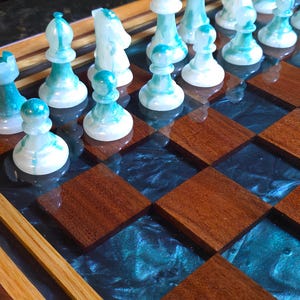 May include: A close-up of a handcrafted chess set. The chess pieces are white with teal accents. The chessboard features alternating squares of dark wood and a teal, resin-like material. The board is framed with light wood.