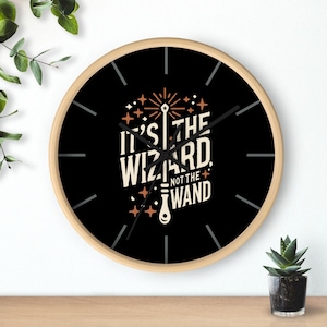 May include: Black and white wall clock with a wooden frame. The clock face features a stylised wand with sparks and the text "It's the wizard, not the wand".