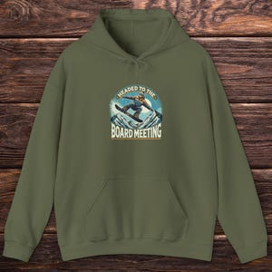 Puede incluir: Una sudadera con capucha verde con un gráfico de un snowboarder y el texto "Headed to the Board Meeting".