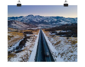 May include: An aerial photograph of a long, straight road leading towards snow-covered mountains. The road is bordered by fields and patches of snow. A vehicle is seen on the road, and the sky displays a gradient of blue and orange.