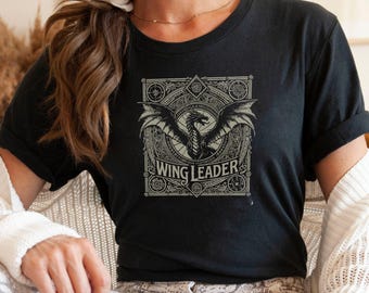 Peut inclure: T-shirt noir avec un graphique de style vintage d'un dragon aux ailes déployées. Le graphique est entouré d'un cadre orné avec le texte "Wing Leader".