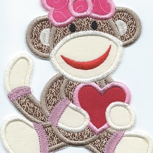 May include: A brown and white monkey with a pink bow holding a red heart. The monkey is wearing a pink and brown knitted sweater.