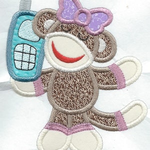 May include: A brown sock monkey with a purple bow, holding a blue toy phone. The monkey has a red smile and pink accents on its arms and feet.