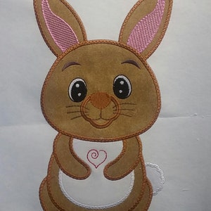 May include: Embroidered brown bunny with pink inner ears, large black eyes, and a white apron featuring a red heart. The bunny has a small white tail and is likely a decorative item or applique.