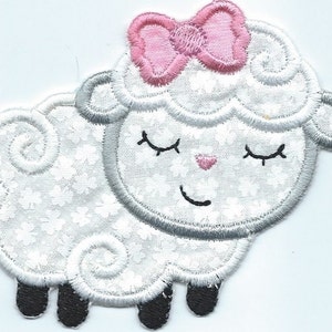 May include: Embroidered patch of a smiling sheep with closed eyes and a pink bow. The sheep is white with a floral pattern, gray trim, and black hooves. A small pink heart is on the sheep's nose.
