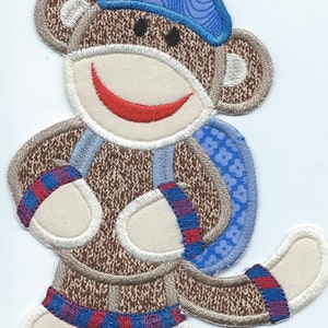 May include: Embroidered sock monkey patch with a blue hat and backpack. The monkey has a smiling red mouth, tan face, and brown body. The patch is designed with a white border and colorful accents.