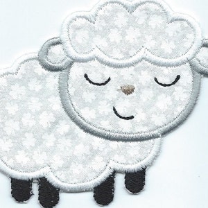 May include: A white and gray embroidered sheep patch with a smiling face and closed eyes. The sheep has a fluffy body with a floral pattern, gray outline, and black legs. The patch is designed for sewing or ironing onto fabric.