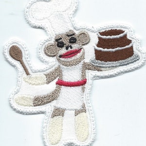 May include: Embroidered patch featuring a sock monkey chef holding a chocolate cake and a wooden spoon. The monkey wears a white chef's hat, apron with a red belt, and has a smiling expression. The patch has a white border.