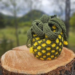 May include: A crocheted pineapple-shaped pouch with a green top and yellow body. The pouch is sitting on a wooden log.