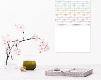 May include: A wall-mounted year planner with a colorful grid design hangs above a table. A vase with pink flowers and a glass with green liquid sit on the table. A gray tray with compartments and a small tool is also on the table.