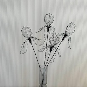 May include: A display of wire flowers in a grey glass vase. The flowers feature intricate, open petals and dark stems. The vase is cylindrical with vertical grooves. The background is a white wall with vertical lines.
