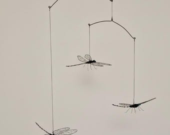 May include: A hanging mobile featuring three dragonfly sculptures. The dragonflies are black with delicate, wire-frame wings. The mobile is suspended from a curved, black metal frame, creating a whimsical and airy decorative piece.