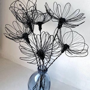 K&ouml;nnte beinhalten: Ein Strau&szlig; schwarzer Drahtblumen in einer blauen Glasvase. Die Blumen haben zarte, offene Bl&uuml;tenbl&auml;tter und sind so angeordnet, dass sie eine volle, k&uuml;nstlerische Darstellung ergeben. Die Vase hat eine weiche, abgerundete Form.