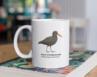 May include: White ceramic mug featuring a detailed illustration of a Black Oystercatcher bird with an orange beak and gray plumage. The mug has the bird's name and scientific name printed below the image. The mug is sitting on a book.