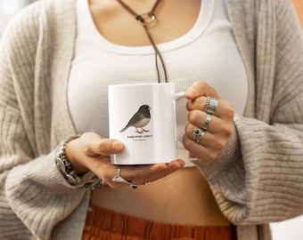 May include: White ceramic mug featuring a Dark-Eyed Junco bird illustration. The bird is depicted in shades of gray and black. The mug has a handle and the text "DARK-EYED JUNCO" is printed below the bird image. A simple, elegant design.