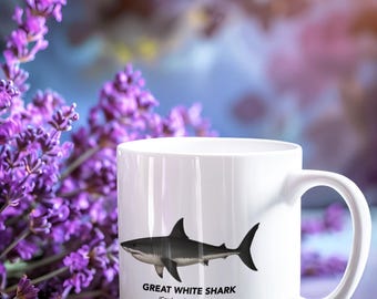May include: A white ceramic mug featuring a black and grey illustration of a great white shark. Below the shark, the text reads GREAT WHITE SHARK and (Carcharodon carcharias) in black capital and lowercase letters respectively.