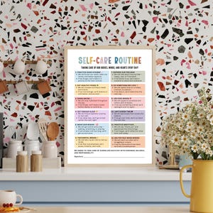May include: A framed print titled "Self-Care Routine" with a beige wooden frame. The print features a list of self-care practices in a colorful design. The background is a terrazzo pattern with a yellow vase of white flowers.