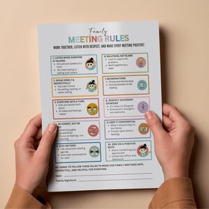 Op de afbeelding: Een bedrukt vel met de titel "Familie Vergaderregels" met kleurrijke illustraties en opsommingstekens. De regels omvatten luisteren, vriendelijk spreken, om de beurt en meningen respecteren. Het document wordt door een persoon vastgehouden.