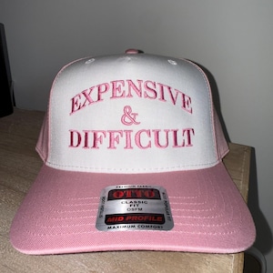 Peut inclure: Casquette de baseball rose et blanche avec les mots "EXPENSIVE & DIFFICULT" brodés en rose. La casquette a une visière incurvée et une étiquette indiquant "OTTO CLASSIC FIT OSFM MID PROFILE MAXIMUM COMFORT".