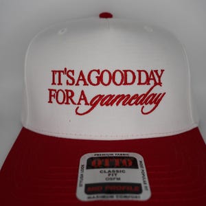 Op de afbeelding: Een witte honkbalpet met een rode klep en een rode knoop aan de bovenkant. De voorkant is voorzien van rood geborduurde tekst die luidt IT'S A GOOD DAY FOR A gameday. Een rechthoekige sticker op de klep toont de merknaam OTTO en CLASSIC FIT.