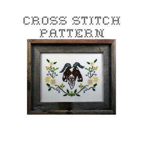 May include: Cross stitch pattern of a brown and white goat's head with yellow and blue flowers in a wreath around it. The pattern is framed in a rustic wooden frame.
