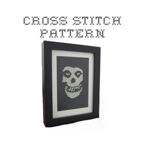 May include: A black framed cross stitch pattern of a white Misfits skull logo. The skull has a wide grin and two large eyes.