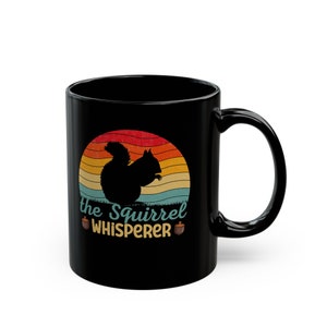 May include: Black coffee mug with a retro-style design featuring a silhouette of a squirrel in front of a sunset. The text "The Squirrel Whisperer" is printed below the squirrel with two acorn illustrations.