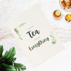 Può includere: Un asciugamano da tè bianco con disegni di foglie verdi e il testo "Una tazza di tè risolve tutto".