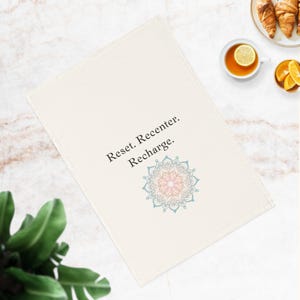 Könnte beinhalten: Ein weißes Leinen-Geschirrtuch mit dem Text "Reset. Recenter. Recharge." und einem blauen und rosafarbenen Mandala-Design.
