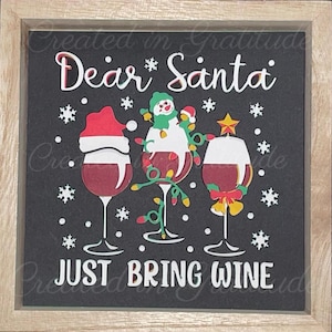 May include: A black framed sign with the text "Dear Santa Just Bring Wine" in white. The sign features a snowman, a Santa hat, and three wine glasses with red wine, all decorated with Christmas lights.