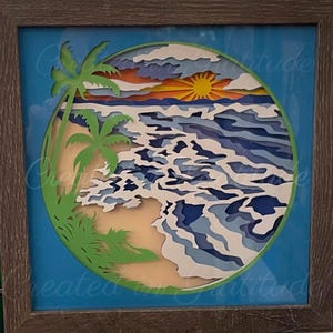 May include: A paper cut-out art piece depicting a beach scene with palm trees, waves, and a sunset. The artwork is framed in a brown wood frame.