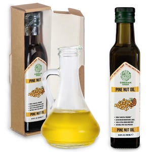May include: A clear glass cruet filled with yellow liquid next to a bottle of pine nut oil. The bottle has a green cap and a label with text and a pine nut illustration. The bottle is in a cardboard box.