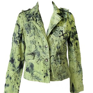 May include: A green denim jacket with black splatter paint and a silver cross detail. The jacket has a button closure and two pockets.