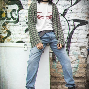 May include: A woman wearing a white t-shirt with a red skeleton hand graphic, a green plaid shirt, blue jeans, and black combat boots. She is standing in front of a brick wall with graffiti.