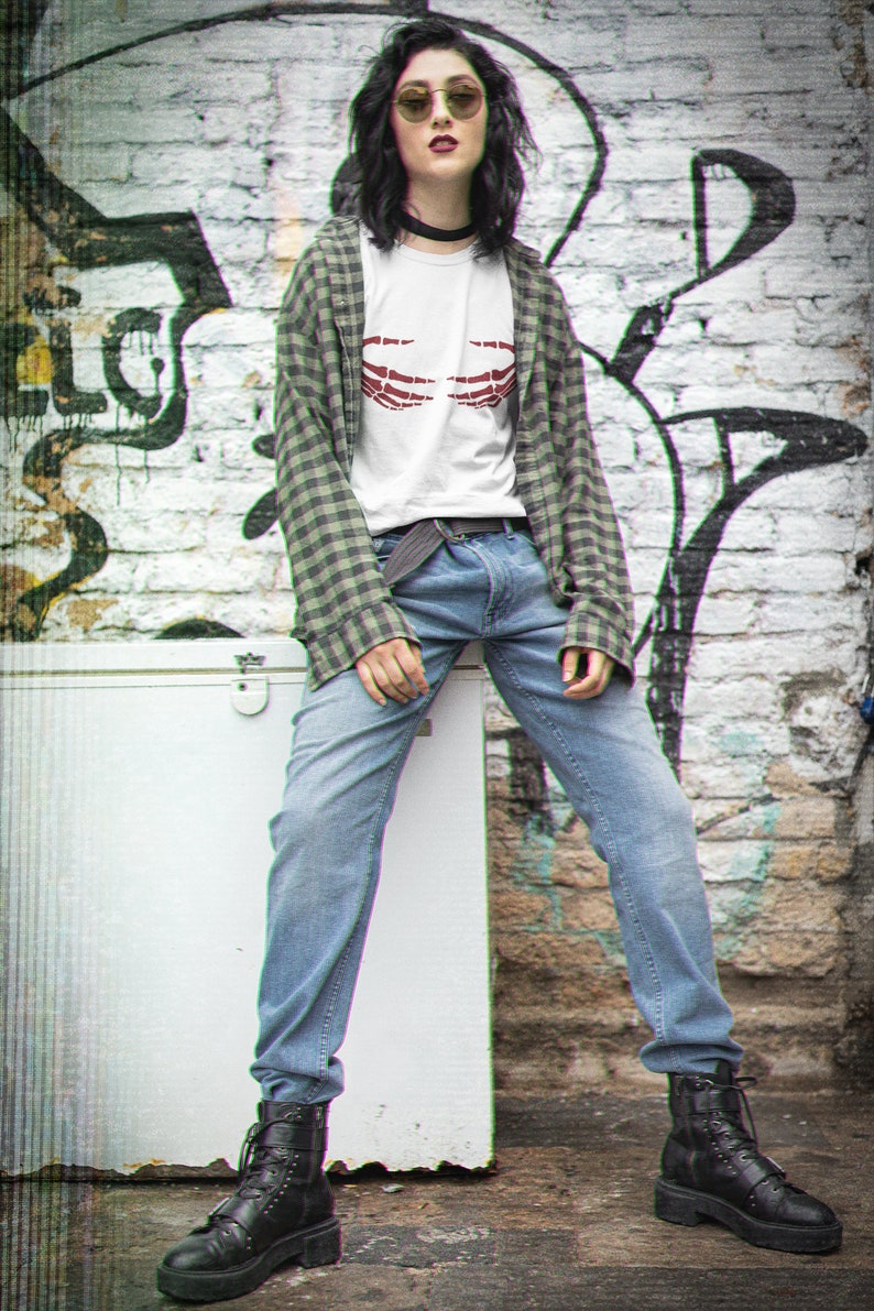 May include: A woman wearing a white t-shirt with a red skeleton hand graphic, a green plaid shirt, blue jeans, and black combat boots. She is standing in front of a brick wall with graffiti.