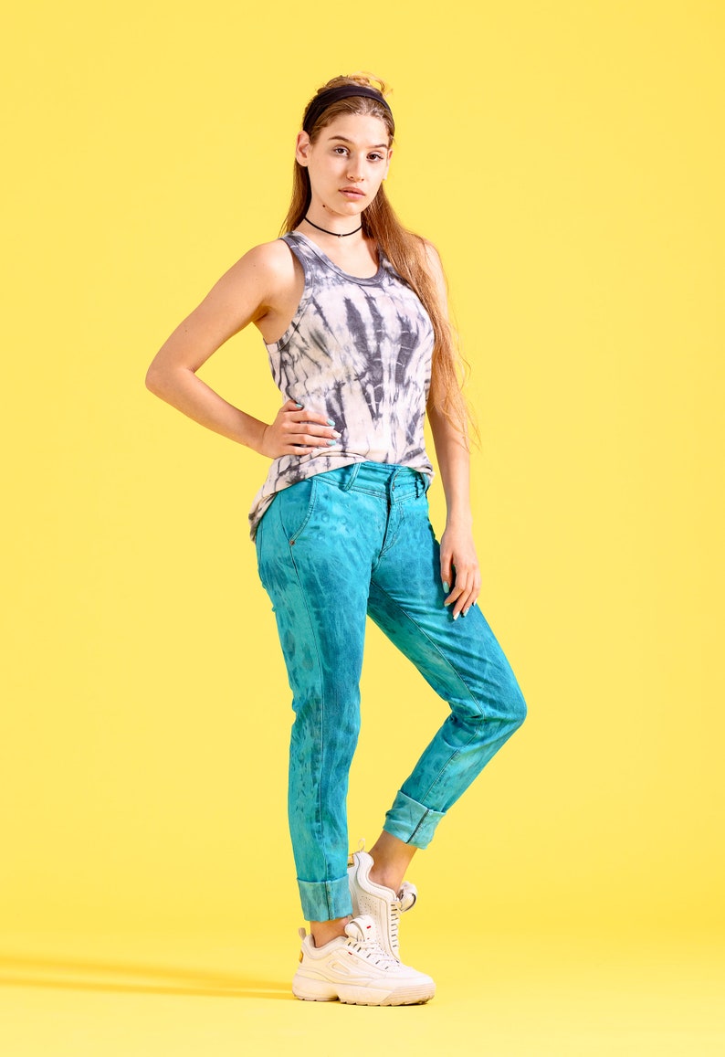 May include: A woman wearing a tie-dye tank top and blue jeans with a white headband. She is standing on a yellow background.