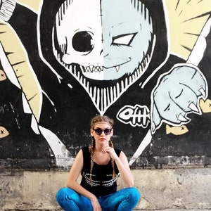 May include: A woman wearing blue jeans and a black tank top with a white skull graphic, poses in front of a black and white mural of a skull with cat ears. She is wearing sunglasses and has blonde hair.