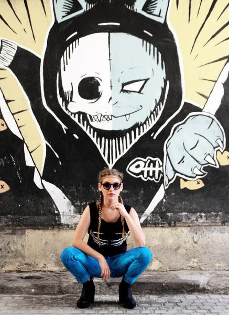 May include: A woman wearing blue jeans and a black tank top with a white skull graphic, poses in front of a black and white mural of a skull with cat ears. She is wearing sunglasses and has blonde hair.