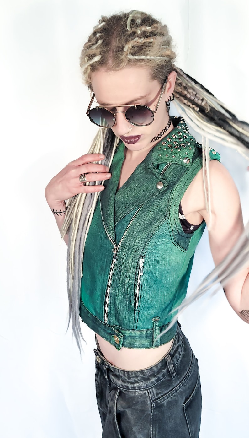 May include: A green denim vest with silver hardware and a studded collar. The vest is cropped and has a zipper closure. The model is wearing the vest over a black shirt and black jeans.