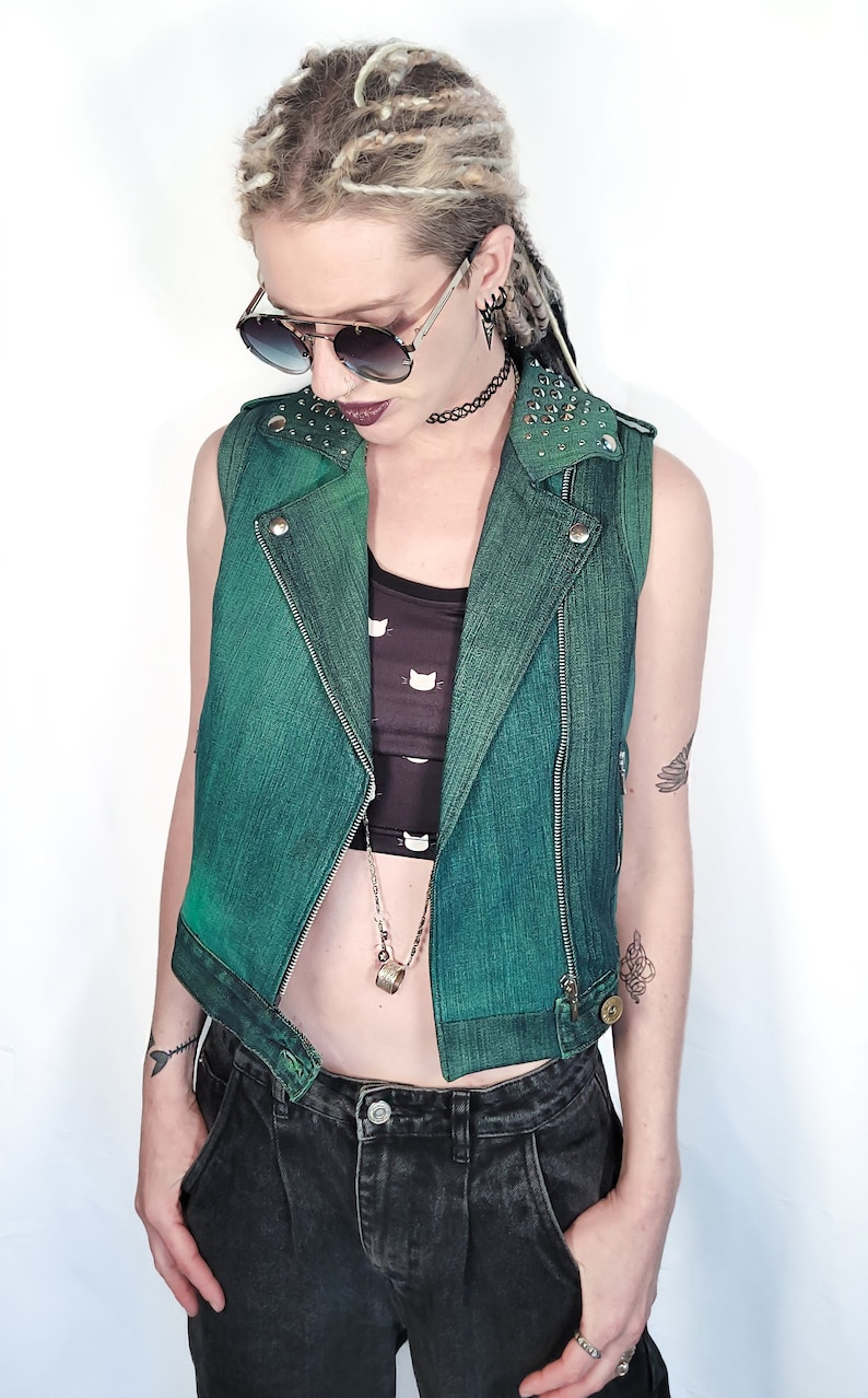 May include: A green denim vest with silver studs on the collar and a zipper closure. The vest is worn over a black tank top with a white cat print. The person is wearing black jeans and has blonde dreadlocks.