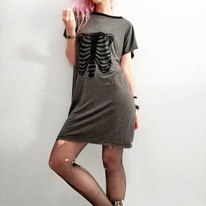 May include: A woman with pink hair is wearing a gray t-shirt dress with a black skeleton graphic. She is also wearing black fishnet tights and leopard print slip-on shoes.