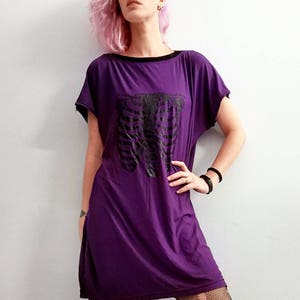 May include: A purple short-sleeved dress with a black ribcage graphic print. The model is wearing black fishnet tights and blue mirrored sunglasses.