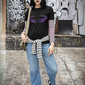 May include: A woman wearing a black t-shirt with a purple skeleton hand graphic, blue jeans, and black and white striped belt. She is wearing black high-top sneakers.