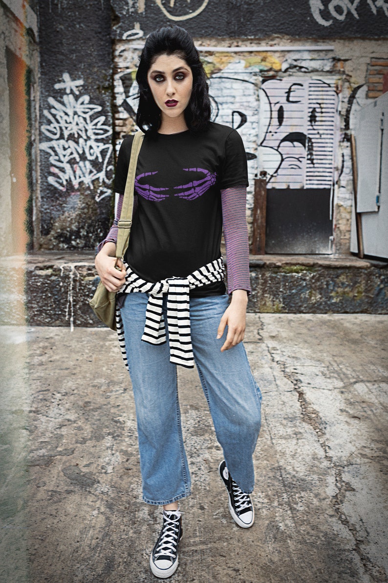 May include: A woman wearing a black t-shirt with a purple skeleton hand graphic, blue jeans, and black and white striped belt. She is wearing black high-top sneakers.